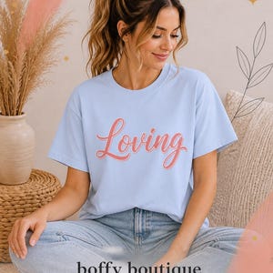 May include: A light blue short-sleeved t-shirt featuring the word Loving printed in a coral-colored script font across the chest. The shirt has a crew neckline and a relaxed fit, paired with light-wash denim jeans and white sneakers.