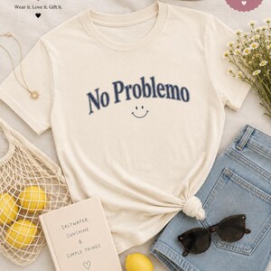 May include: Cream-colored t-shirt with the phrase "No Problemo" in navy blue, featuring a smiling face. The shirt is styled with denim shorts, sunglasses, and a straw hat. A book with the text "Saltwater Sunshine & Simple Things" is also present.
