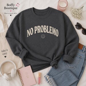 May include: A dark gray sweatshirt with the text "NO PROBLEMO" in a cream-colored, arched font, and a small smiley face. The sweatshirt is styled with blue jeans, headphones, and a tote bag.
