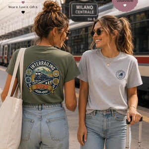 May include: An olive green t-shirt featuring a large circular graphic on the back with a train, mountains, and the text "INTERRAILING 2026" and a light gray t-shirt with a small circular "EUROPE 2026" logo on the front left chest area.