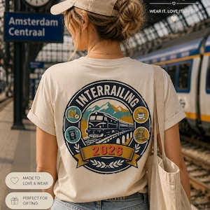 May include: A beige t-shirt featuring a circular graphic on the back with a train, mountains, and the text "INTERRAILING 2026". The design includes small icons of a map, a passport, a coffee cup, and a backpack. The shirt is worn with blue jeans.