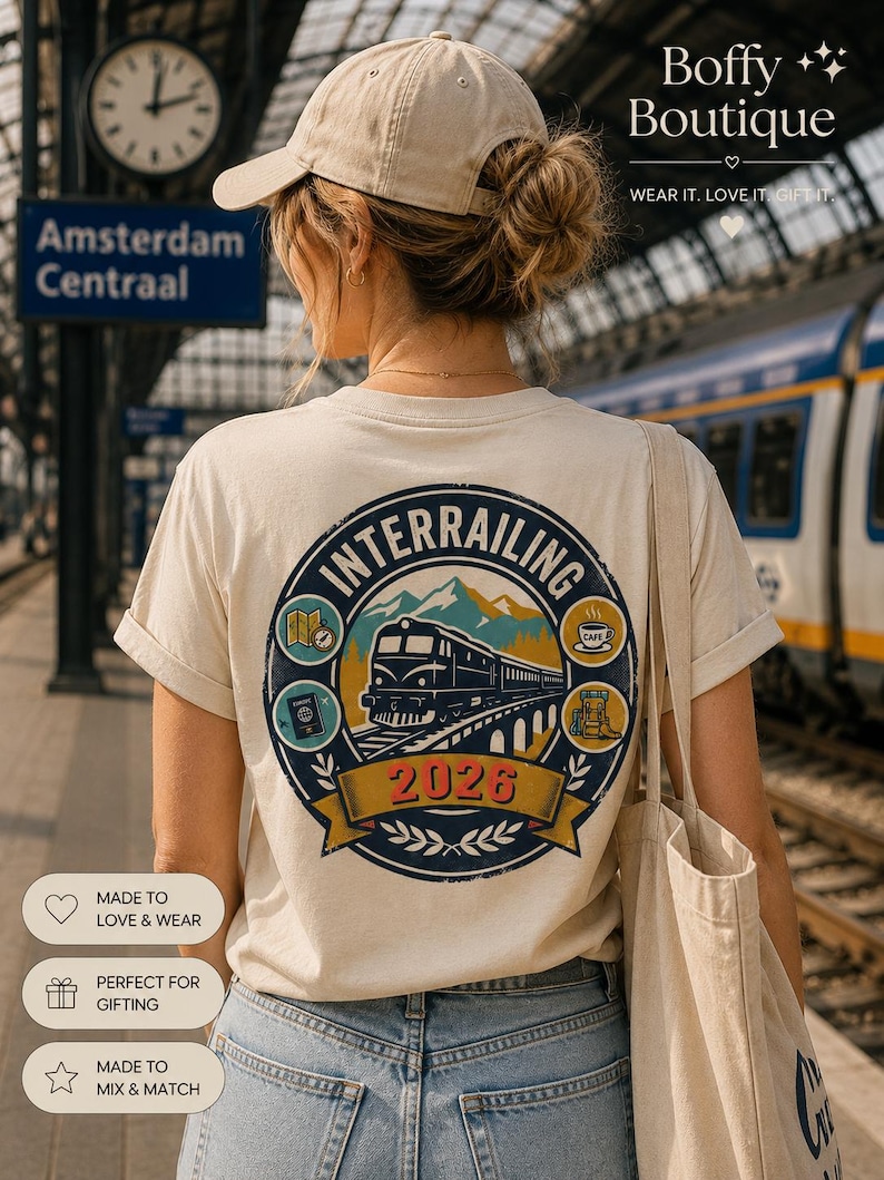 May include: A beige t-shirt featuring a circular graphic on the back with a train, mountains, and the text "INTERRAILING 2026". The design includes small icons of a map, a passport, a coffee cup, and a backpack. The shirt is worn with blue jeans.