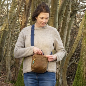 May include: A brown crossbody bag with a blue strap and a yellow zipper. The bag is worn over a light brown sweater and blue jeans. The background features a forest with tall trees.