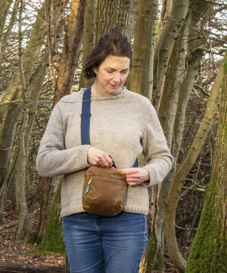 May include: A brown crossbody bag with a blue strap and a yellow zipper. The bag is worn over a light brown sweater and blue jeans. The background features a forest with tall trees.
