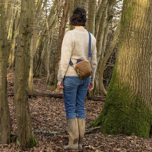 May include: A person wearing a tan sweater, blue jeans, and brown boots stands in a forest. A brown crossbody bag with a blue strap is worn across the body. The forest floor is covered in fallen leaves, and trees surround the person.