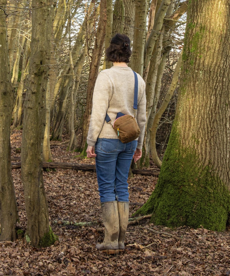 May include: A person wearing a tan sweater, blue jeans, and brown boots stands in a forest. A brown crossbody bag with a blue strap is worn across the body. The forest floor is covered in fallen leaves, and trees surround the person.