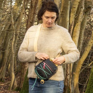 May include: A black crossbody bag with a cream-colored strap and a colorful design. The bag has a rectangular shape with a zipper closure and a gold-colored metal D-ring. The bag is worn over the shoulder.