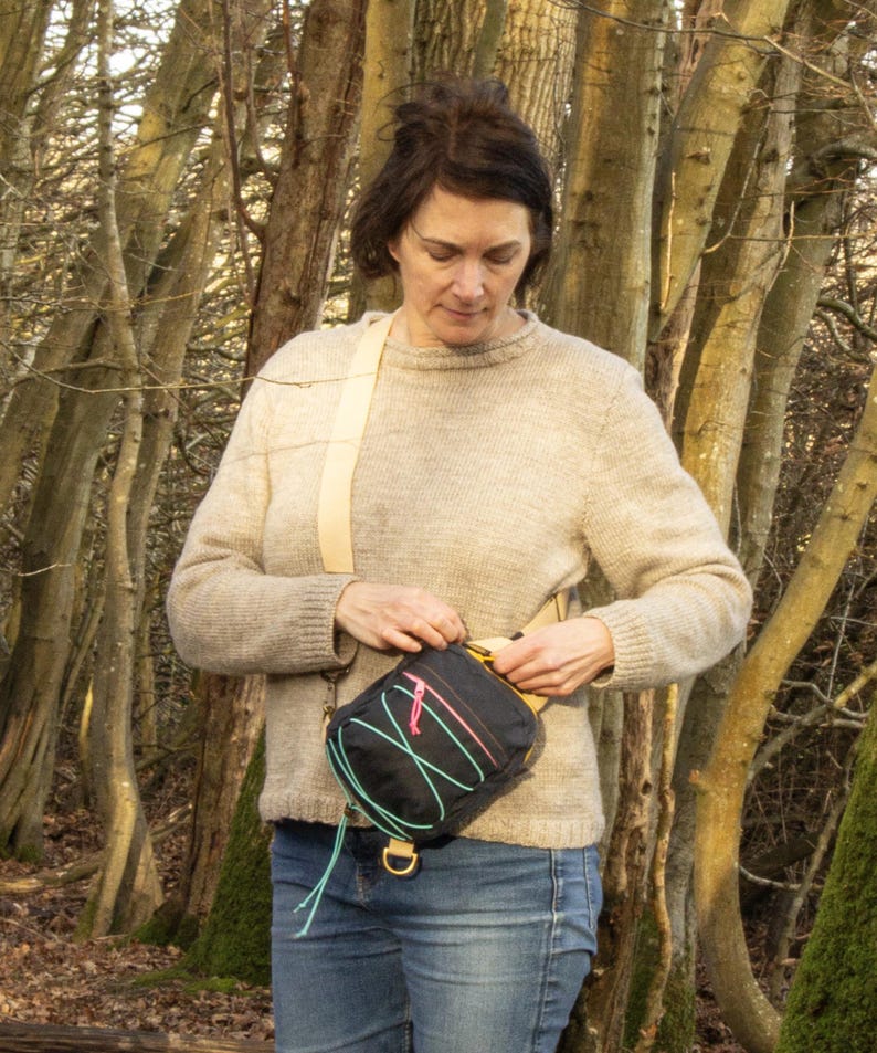 May include: A black crossbody bag with a cream-colored strap and a colorful design. The bag has a rectangular shape with a zipper closure and a gold-colored metal D-ring. The bag is worn over the shoulder.