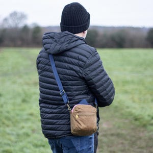 May include: A person wearing a black quilted jacket, black beanie, and blue jeans. A brown crossbody bag with a pink zipper and blue strap is worn over the shoulder. The background is a green field and overcast sky.