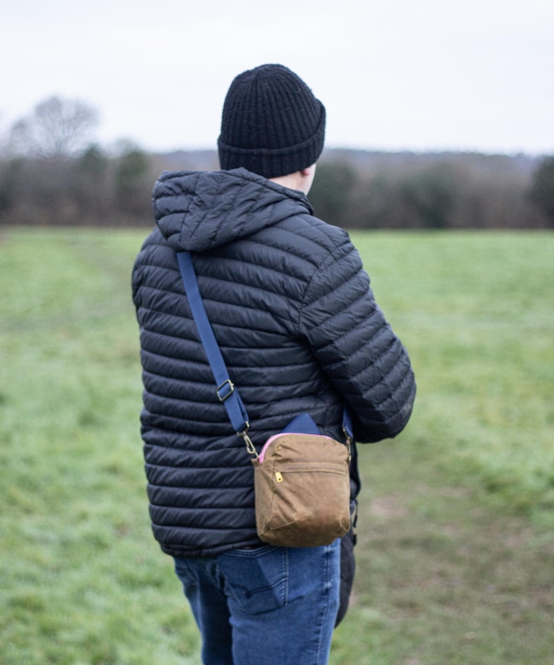 May include: A person wearing a black quilted jacket, black beanie, and blue jeans. A brown crossbody bag with a pink zipper and blue strap is worn over the shoulder. The background is a green field and overcast sky.