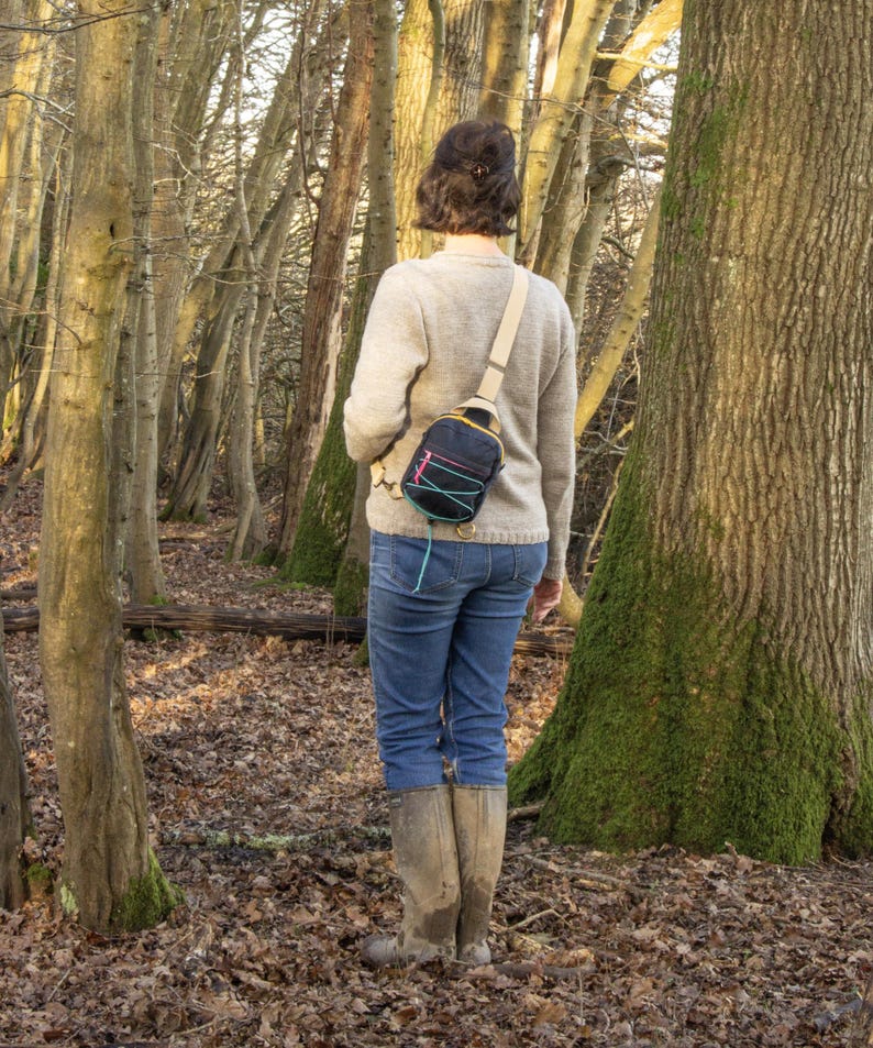 May include: A person wearing a light gray sweater, blue jeans, and tall rubber boots stands in a forest. A small black and yellow sling bag with teal and pink accents is worn across the body.