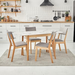 May include: A rectangular dining table with a white marble-patterned top and light wood legs, accompanied by four chairs featuring light wood frames and grey upholstered seats, arranged on a grey area rug in a kitchen setting.