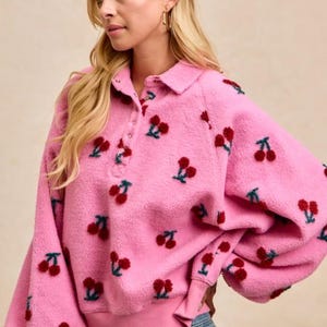 Pode incluir: Camisola rosa de lã polar com gola e bordado de cerejas. A camisola de manga comprida tem uma carcela de botões e punhos canelados. As cerejas são vermelhas com caules verdes.