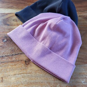 Könnte beinhalten: Zwei gerippte Strickmützen, eine in Staubrosa und die andere in Dunkelmarineblau. Die Mützen haben umgeschlagene Krempen und liegen auf einer Holzoberfläche. Dies sind Mützen im lässigen Stil.