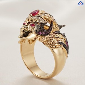 Puede incluir: Un anillo de oro con un rostro esculpido, una piedra preciosa roja de talla marquesa en la frente, dos piedras rojas más pequeñas como ojos y diamantes blancos y negros engastados en pavé a lo largo de la base del diseño metálico.