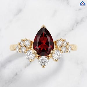 Op de afbeelding: Een gouden ring met een centrale peervormige, dieprode granaat edelsteen. De ring is versierd met een cluster van kleine, ronde, heldere witte diamanten die symmetrisch aan beide zijden van de primaire rode steen zijn geplaatst.