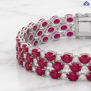 Könnte beinhalten: Ein mehrreihiges Armband aus silberfarbenem Metall mit zahlreichen oval geschliffenen, leuchtend roten Rubinen in drei Reihen. Zwischen den Rubinen sind kleine, klare, rund geschliffene weiße Steine eingefasst, ergänzt durch einen Kastenschloss.