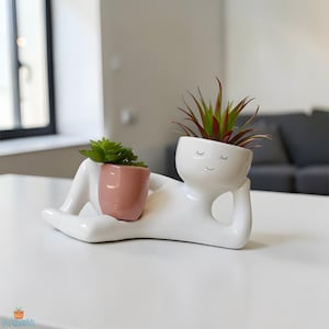 Pode incluir: Um vaso de cerâmica branca em forma de pessoa reclinada, segurando um pequeno vaso rosa e um vaso branco maior com um rosto sorridente. Os vasos contêm pequenas plantas verdes e de pontas vermelhas. O vaso está sobre uma mesa branca.