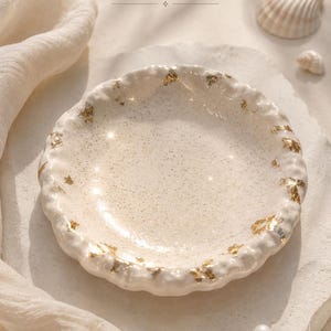 Peut inclure: Un plat en céramique crème avec un bord texturé et des accents de feuilles d'or. Le plat rond est posé sur une surface claire, avec le texte "ÉCUME & ÉCLAT" au-dessus. Des coquillages et une perle sont également visibles.