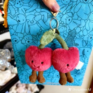 Op de afbeelding: Een pluche kersen sleutelhanger met een lachend gezicht, bruine benen en een groene stengel en blad. De sleutelhanger is bevestigd aan een blauwe tas met een herhalend patroon van cartoon dieren.