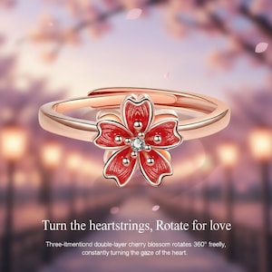 May include: Rose gold adjustable ring featuring a red cherry blossom flower with a central clear gemstone. The flower has red enamel petals and small gold accents. The ring's design includes the text "Turn the heartstrings, Rotate for love."