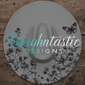 May include: A grey fedora hat with a floral and butterfly design. The hat features the text "Vaughtastic Designs" in a teal and white font. The brim is decorated with black and white flowers and a butterfly.