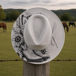 May include: A light gray felt fedora hat featuring a black and white ink-style illustration of a cowboy boot surrounded by sunflowers on the brim. The hat includes a matching thin gray cord tied in a simple knot on the side of the crown.