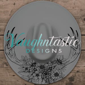 Könnte beinhalten: Ein grauer Fedora-Hut mit breiter Krempe. Der Hut hat ein Blumen- und Geweih-Design in Schwarz und Weiß, mit dem Text "Vaughntastic DESIGNS" in Türkis und Weiß auf dem Oberteil. Ein modisches Accessoire.