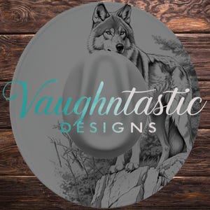May include: A gray cowboy hat featuring a detailed illustration of a wolf standing on a rock. The hat has the text "Vaughntastic DESIGNS" in teal and white. The background is a wooden surface.