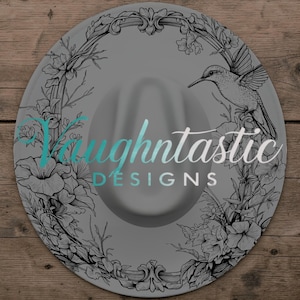 May include: A round gray hat featuring a black line art illustration of a hummingbird surrounded by floral vines and an ornate frame. The text Vaughtastic DESIGNS is printed in a teal and white script and sans-serif font across the center.