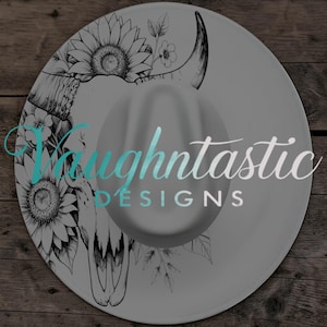 May include: A gray fedora-style hat with a decorative black and white floral and bull skull design. The hat features the text "Vaughtnastic DESIGNS" in a gradient teal and silver color.