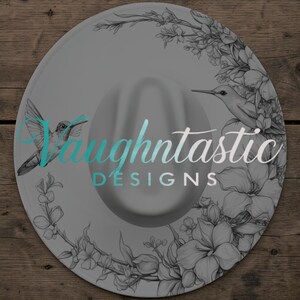 May include: A round, light gray hat featuring a black and white line art illustration of hummingbirds and floral vines. The text "Vaughntastic DESIGNS" is printed in a teal and white script and sans-serif font across the center of the hat.
