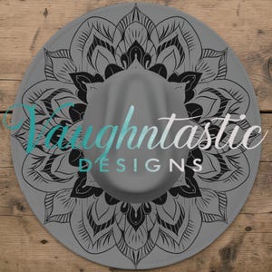 Könnte beinhalten: Ein grauer Fedora-Hut mit einem schwarzen Mandala-Design auf der Krempe. Der Hut trägt den Text "Taughtnastic DESIGNS" in Türkis und Weiß. Der Hut liegt auf einem Holzgrund.