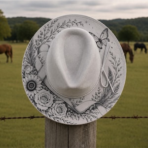 Op de afbeelding: Een lichtgrijze fedora-hoed met een gedetailleerde zwarte inktillustratie van een vlinder, zonnebloemen, rozen en diverse wilde bloemen. Een bijpassende lichtgrijze lintband zit rond de kroon en is aan de rechterkant geknoopt.