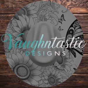 May include: A gray wide-brimmed hat featuring a black line art illustration of sunflowers and butterflies across the crown and brim. The text "Vaughntastic DESIGNS" is printed in a teal and white script font across the center of the hat.