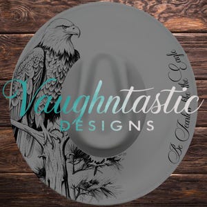 Könnte beinhalten: Ein grauer Fedora-Hut mit einer schwarzen Strichzeichnung eines Weißkopfseeadlers, der auf einem Ast sitzt. Der Text Vaughtastic DESIGNS ist in der Mitte aufgedruckt und der Satz Be fearless like the eagle steht an der Seite des Hutes.