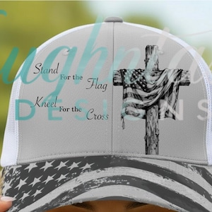 May include: A grey and white trucker hat featuring a black and white graphic of a cross draped with an American flag. The text reads Stand for the Flag, Kneel for the Cross. The brim displays a distressed American flag pattern in black and white.