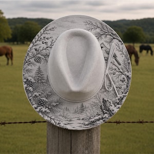 May include: A light grey fedora hat featuring a black and white toile-style print of a winter forest scene with pine trees, snow-covered branches, and a bird. The hat includes a thin, matching light grey cord wrapped around the crown.