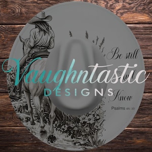 Puede incluir: Un sombrero gris de ala ancha con una ilustración en blanco y negro de una persona con sombrero de vaquero montando a caballo por un campo de flores. El texto dice Be still and know Psalms 46:10, con la marca de agua Vaughtastic DESIGNS.