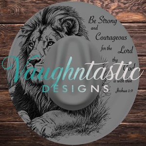 May include: A grey fedora hat featuring a black ink-style illustration of a lion's head and the text Be Strong and Courageous for the Lord thy God is with thee Joshua 1:9. The hat also displays the brand name Vaughtastic DESIGNS in teal.