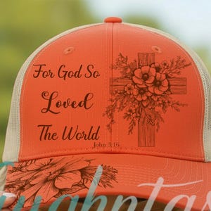May include: An orange trucker hat with a beige mesh back. The front panel features a dark brown graphic of a wooden cross adorned with flowers and the text "For God So Loved The World John 3:16". The brim also displays a floral line art design.
