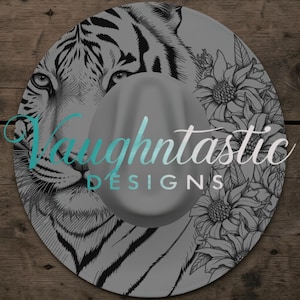May include: A gray fedora hat with a striking design. The hat features a detailed black and white illustration of a tiger's face on the brim, complemented by floral accents. The text "Vaughtntastic DESIGNS" is displayed on the hat.