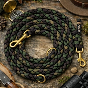 Op de afbeelding: Gevlochten camouflage hondenriem met goudkleurige hardware. De riem is groen, bruin en zwart. De afbeelding bevat ook een kompas, een verrekijker, een hoed en de tekst "Forest Ranger".