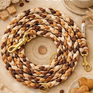Op de afbeelding: Gevlochten hondenriem in bruin-, beige- en crèmetinten met goudkleurige hardware. De riem ligt op een houten oppervlak, met een kopje koffie, koekjes en de tekst "Vanilla Latte" op de achtergrond. De riem heeft een label met "ATLAI".