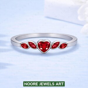 Könnte beinhalten: Ein silberner Ring mit einem herzförmigen roten Edelstein in der Mitte, flankiert von vier kleineren, ovalen roten Edelsteinen. Der Ring ist schmal und poliert. Der Text "NOORE JEWELS ART" befindet sich unten.