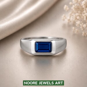 Könnte beinhalten: Ein silberfarbener Ring mit einem rechteckigen, im Smaragdschliff geschliffenen, tiefblauen Edelstein, der in eine glatte, polierte Metallschiene gefasst ist. Das Design zeigt eine klare, geometrische Zargenfassung, die den leuchtend blauen Stein einrahmt.