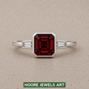 Könnte beinhalten: Ein silberfarbener Ring mit einem großen, rechteckigen, tiefroten Edelstein in einer Zargenfassung, flankiert von zwei kleineren, klaren, spitz zulaufenden Baguette-Steinen auf jeder Seite. Der Text NOORE JEWELS ART ist unten sichtbar.