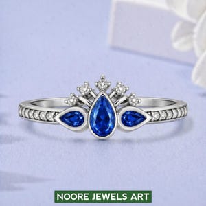 Könnte beinhalten: Ein silberner Ring mit Kronen-Design, mit einem großen, birnenförmigen blauen Edelstein und zwei kleineren blauen Edelsteinen. Das Band ist mit kleinen, klaren Steinen besetzt. Der Text "NOORE JEWELS ART" befindet sich unten.