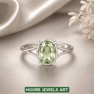 May include: A silver ring with a light green oval gemstone. The ring has a split shank design and four prongs securing the gemstone. The text "NOORE JEWELS ART" is at the bottom of the image.