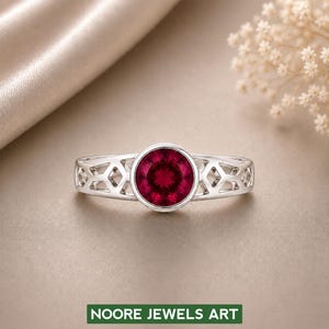 Könnte beinhalten: Ein Silberring mit einem runden, tiefroten Edelstein in einer Zargenfassung. Die Ringschiene weist auf beiden Seiten ein durchbrochenes geometrisches Gittermuster auf. Ein grünes rechteckiges Etikett unten zeigt den weißen Text NOORE JEWELS ART.